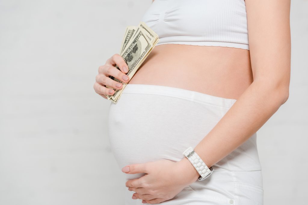 Cropped view of pregnant woman holding dollar banknotes and touching belly on grey background, concept of surrogacy maternity Cropped view of pregnant woman holding dollar banknotes and touching belly on grey background, concept of surrogacy maternity