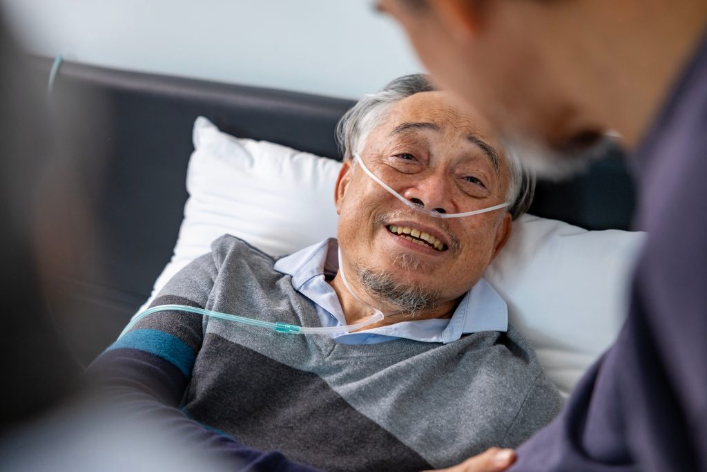 Senior fatal dying male patient with oxygen mask is getting consoling support from his friend and family while suffering from final stage cancer at home with palliative medical treatment concept Senior fatal dying male patient with oxygen mask is getting consoling support from his friend and family while suffering from final stage cancer at home with palliative medical treatment