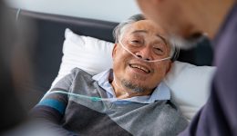 Senior fatal dying male patient with oxygen mask is getting consoling support from his friend and family while suffering from final stage cancer at home with palliative medical treatment