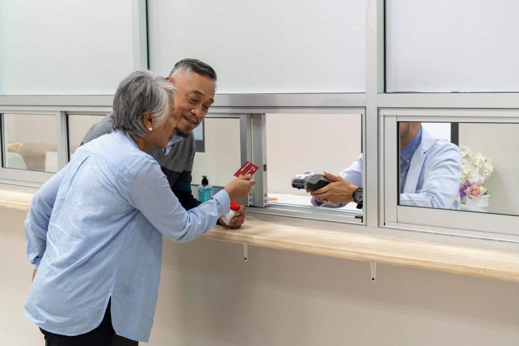 Adult mature couple stand in front of cashier counter of drug department paying with credit card to payment transaction machine at cashier counter in hospital. Pharmacist service or patient counseling