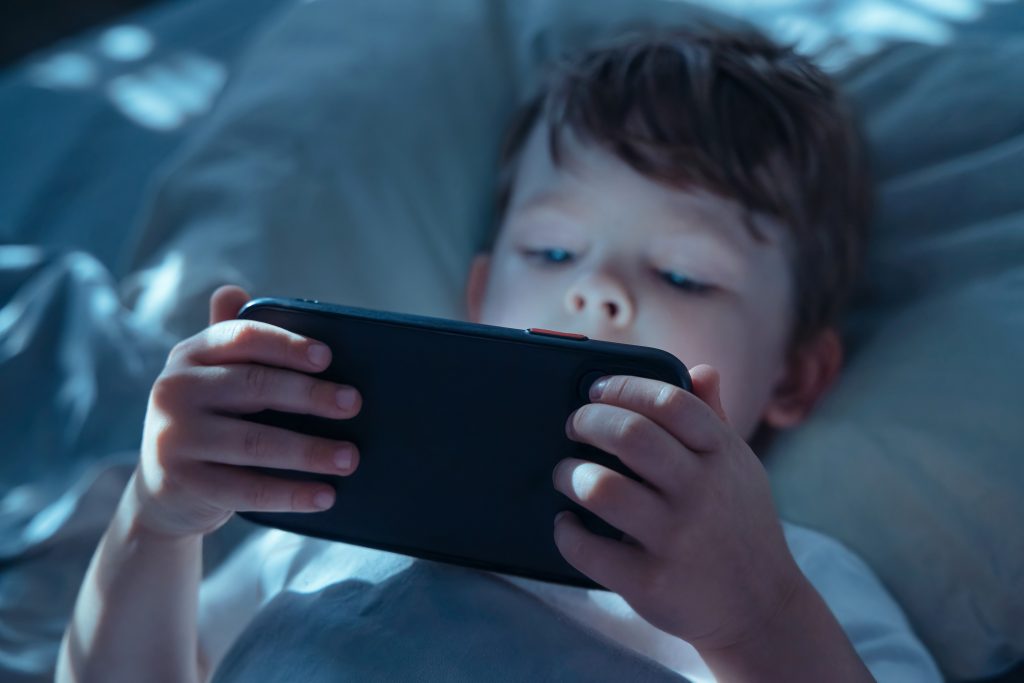 Little child lying in bed in dark room, holding smartphone close and watching intently, illuminated by blue screen light. Gadget addiction and screen dependency. Selective focus on smartphone