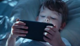 Little child lying in bed in dark room, holding smartphone close and watching intently, illuminated by blue screen light. Gadget addiction and screen dependency. Selective focus on smartphone