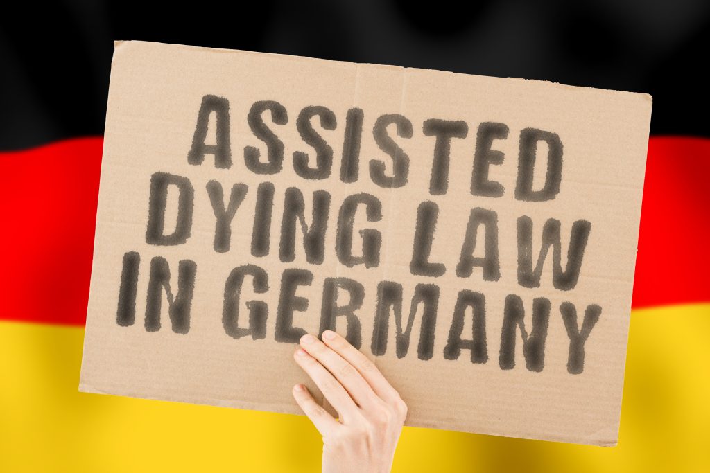 The phrase " Assisted dying law in Germany " on a banner in men's hands with a blurred Deutsch flag in the background. Pain. Care. Clinic. Sickness. Ethical. Ethic. Depression. Drug. End. Judge