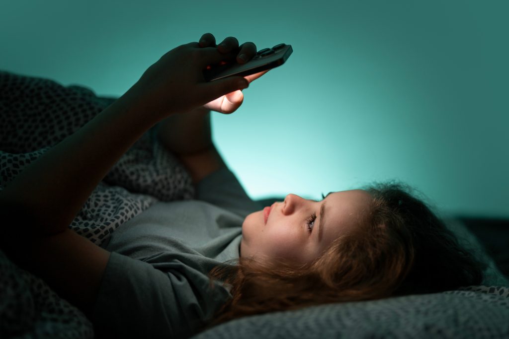 Teenager chatting on phone in bed at night. Child texting on phone in bed at night. Kid scrolling phone under blanket at night. Teenager browsing internet on phone in bed at night